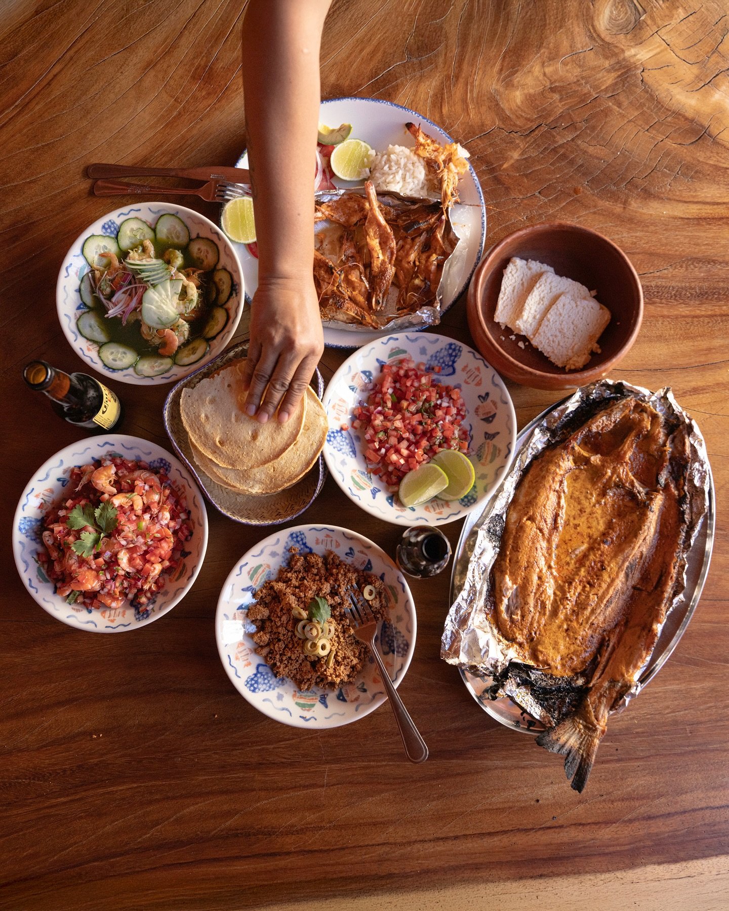 Mesa de madera vista desde arriba con pescado, tortillas y varios platillos para compartir; una mano toma una tortilla.
